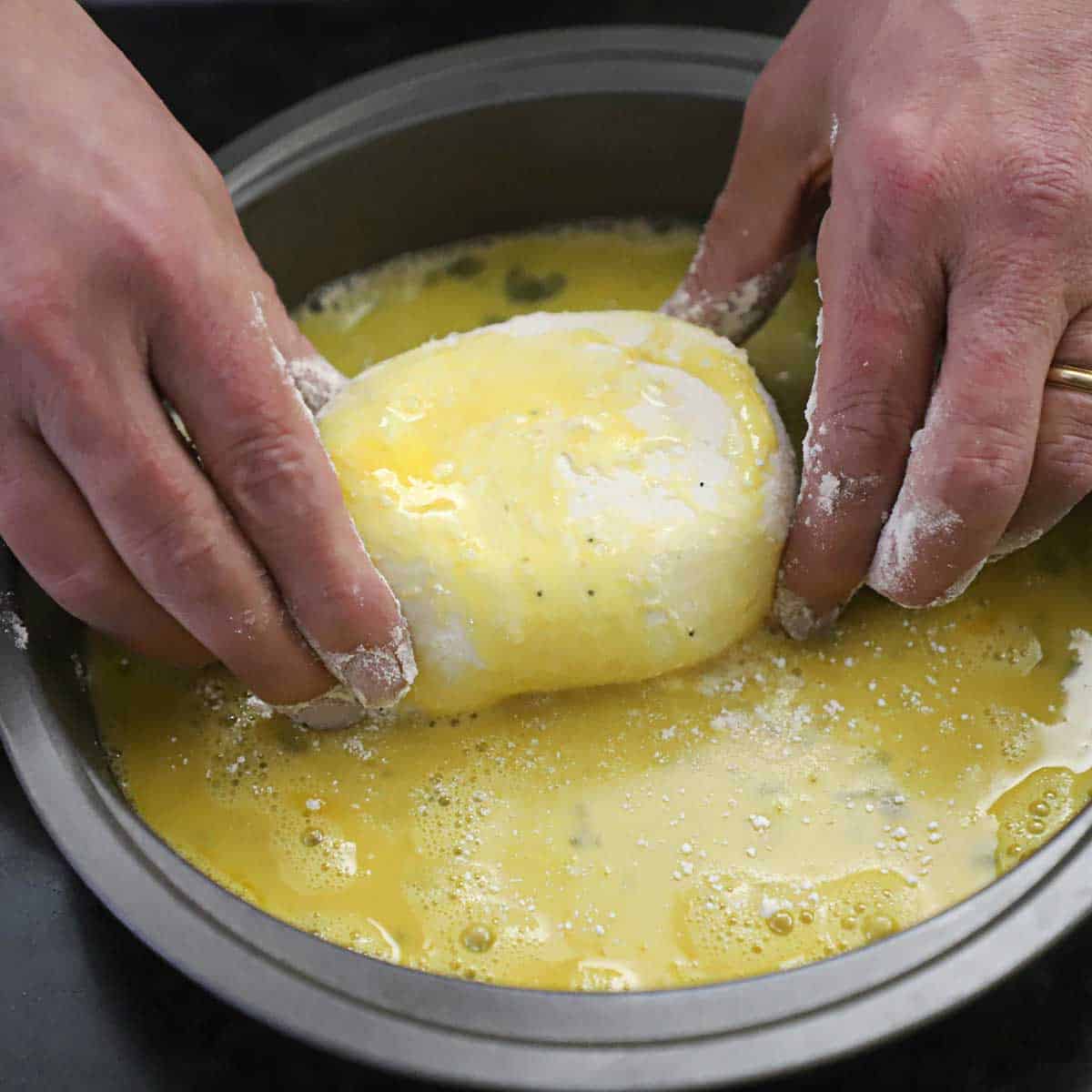 A person using two hands to coat an egg wash all over a large ball of burrata cheese in a baking pan.