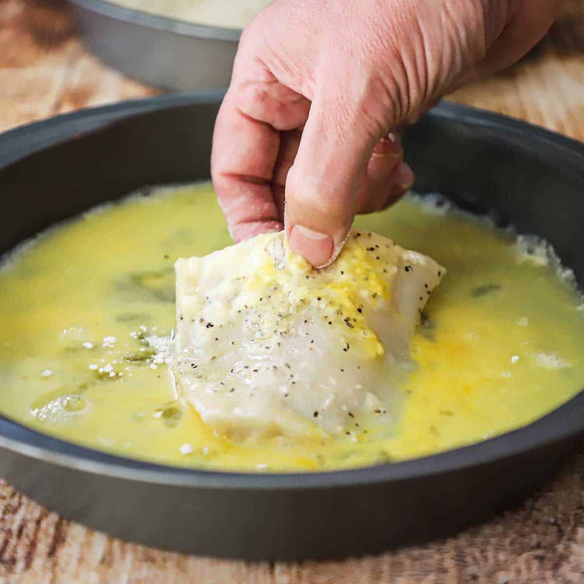 A person submerging a filet of cod that has been coated with seasoned flour in an egg bath in a round baking pan.