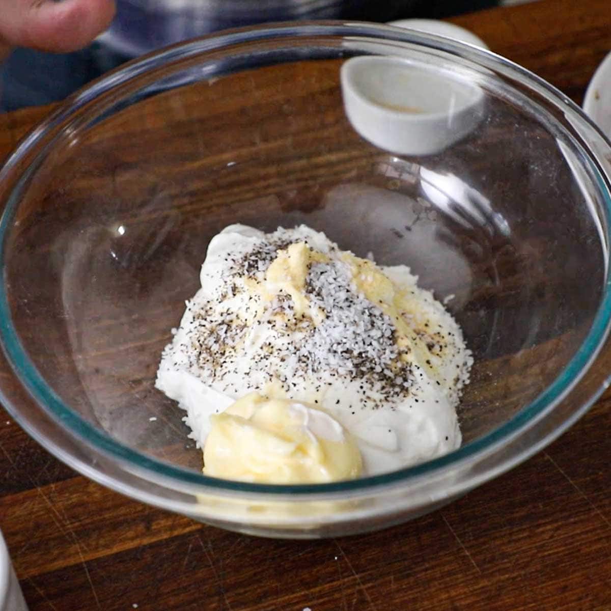 A large glass bowl filled with sour cream, mayonnaise, salt, pepper, and garlic powder resting on a wooden cutting board.
