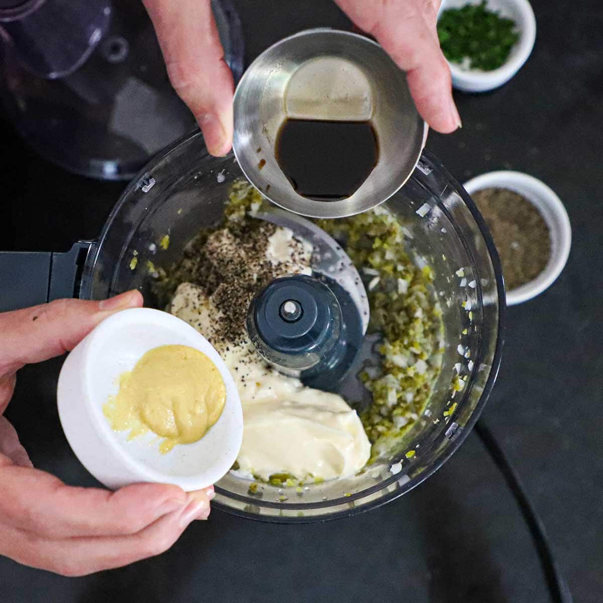 A person getting ready to transfer Worcestershire sauce from a small bowl and Dijon mustard from another bowl into a food processor filled with chopped pickles, onions, mayonnaise, and seasonings.