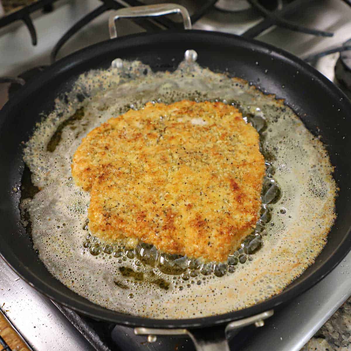 A large, thin breaded chicken cutlet being fried with butter and oil in a large, black skillet on a gas stove.