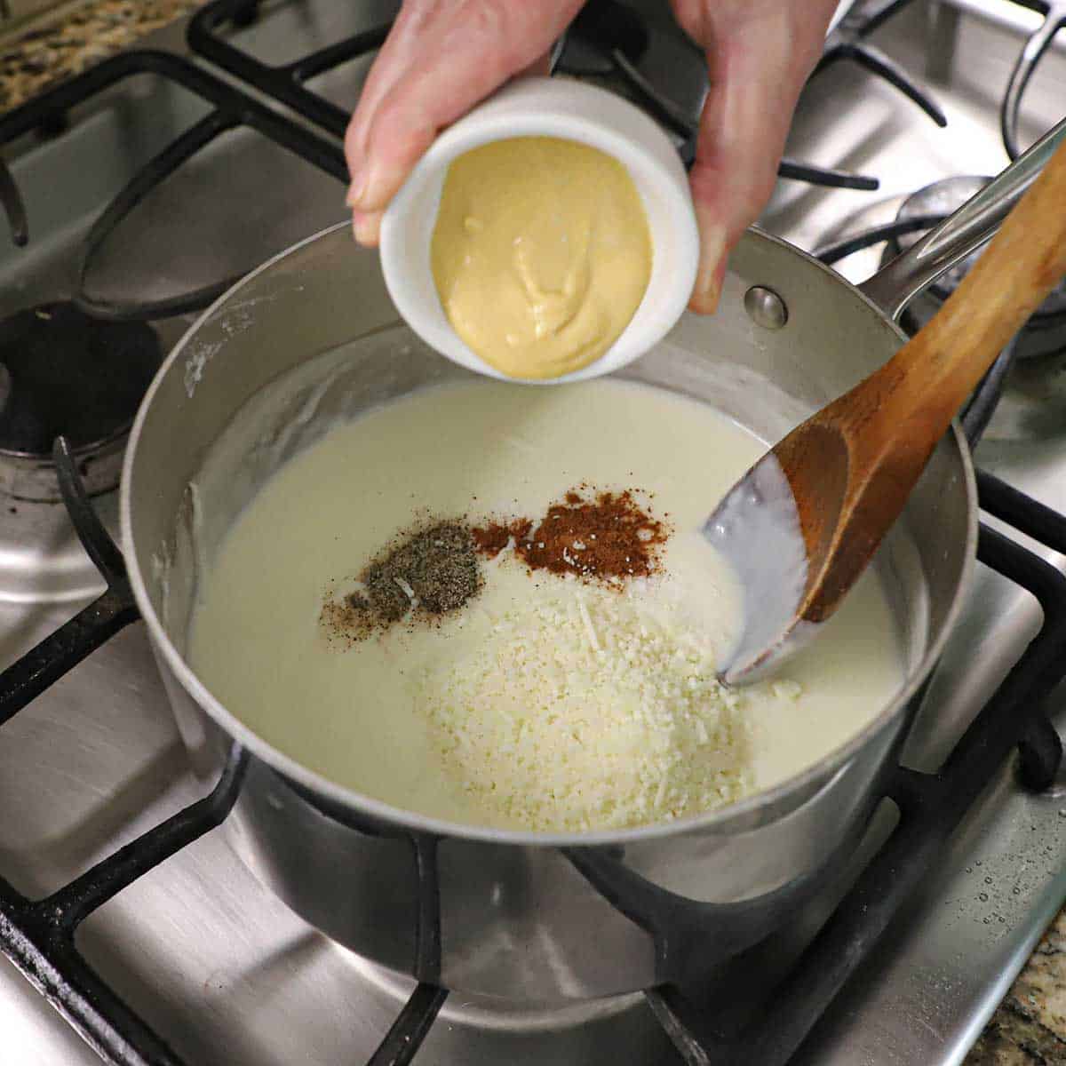 A person transferring Dijon mustard from a small white bowl into a saucepan that is filled with a bechamel sauce with small mounds of pepper, paprika, and grated parmesan on the surface.