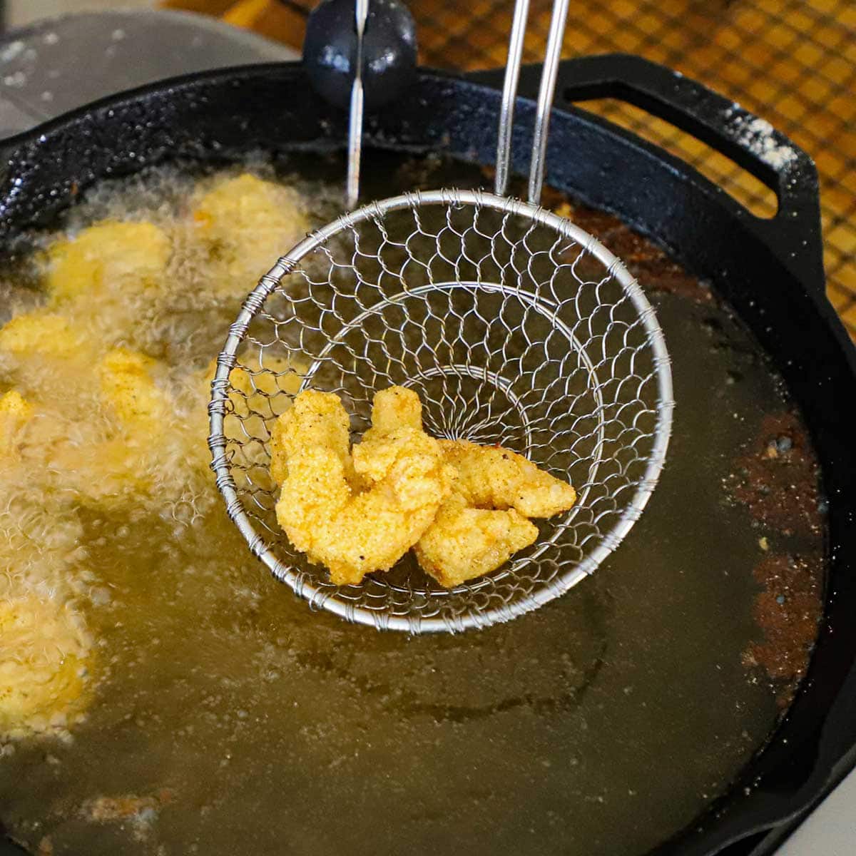 A metal spider being used to raise lightly fried shrimp from a large cast-iron skillet that is filled with hot oil.