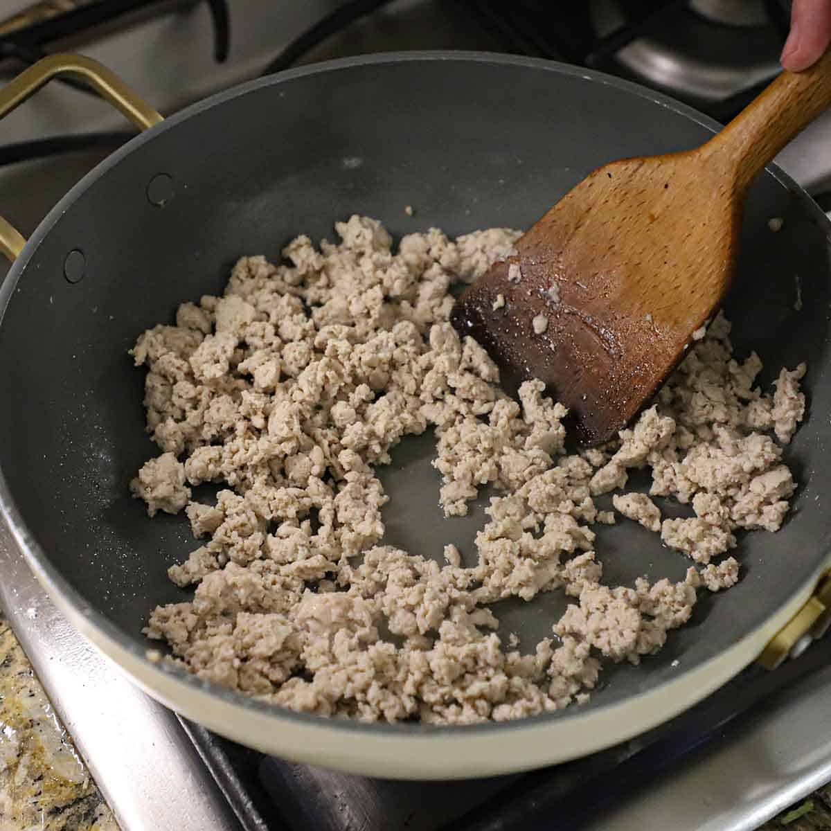 A person using a wooden spatula to stir ground chicken that is being cooked in a large non-stick skillet on a gas stove.