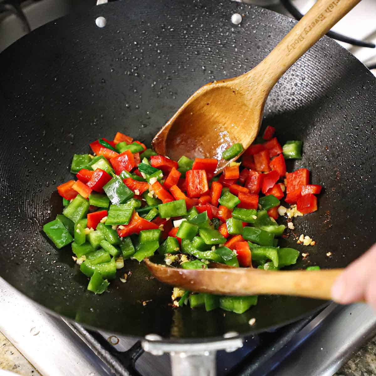 A person using two large wooden spoons to stir-fry roughly chopped red bell pepper, green bell pepper, and minced garlic and ginger in a large wok on a gas stove.