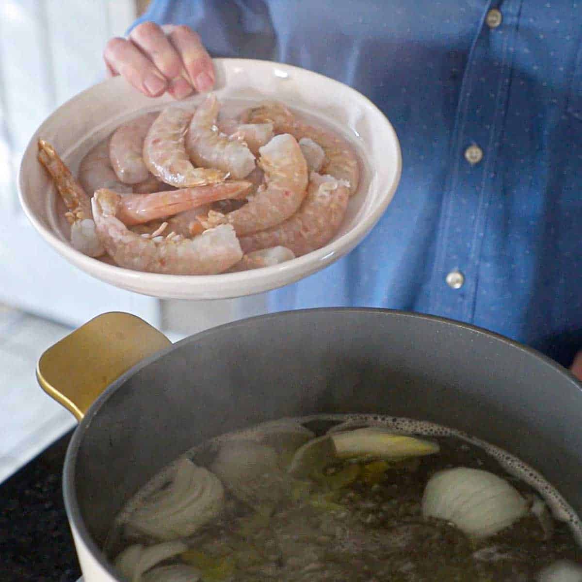 A person holding a shallow white bowl filled with uncooked pink shrimp over a pot of simmering water filled with aromatics.
