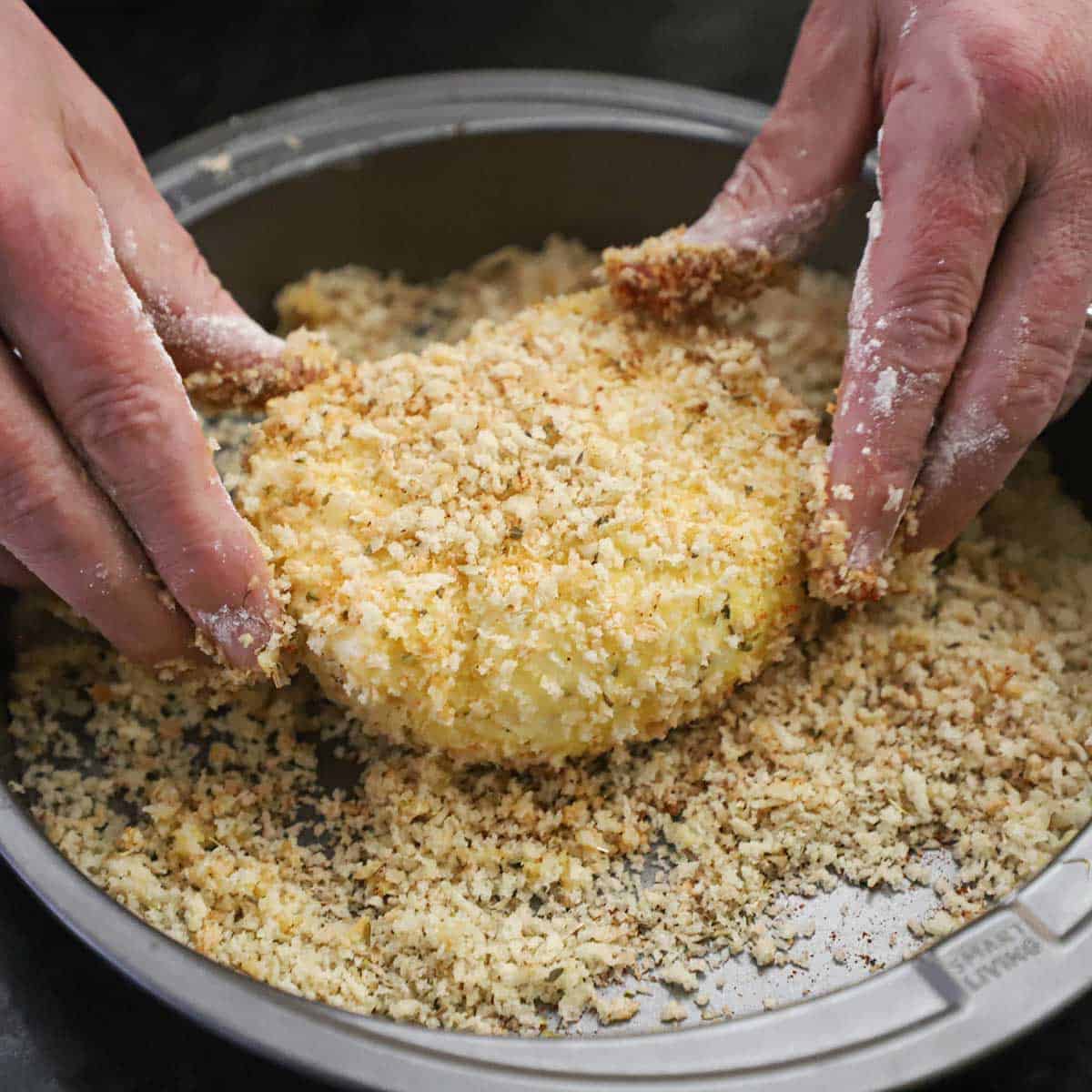 A person using two hands to dredge a large ball of burrata cheese that had ben covered in an egg wash and it now being covered all over with seasoned Panko breadcrumbs.