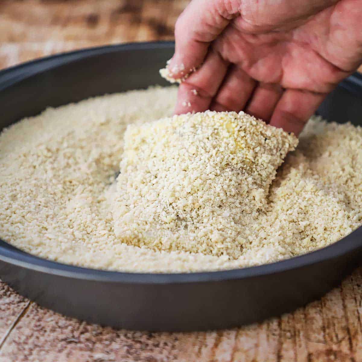 A person dredging a filet of cod that was floured and then coated with egg through Panko breadcrumbs that are in a round baking pan.