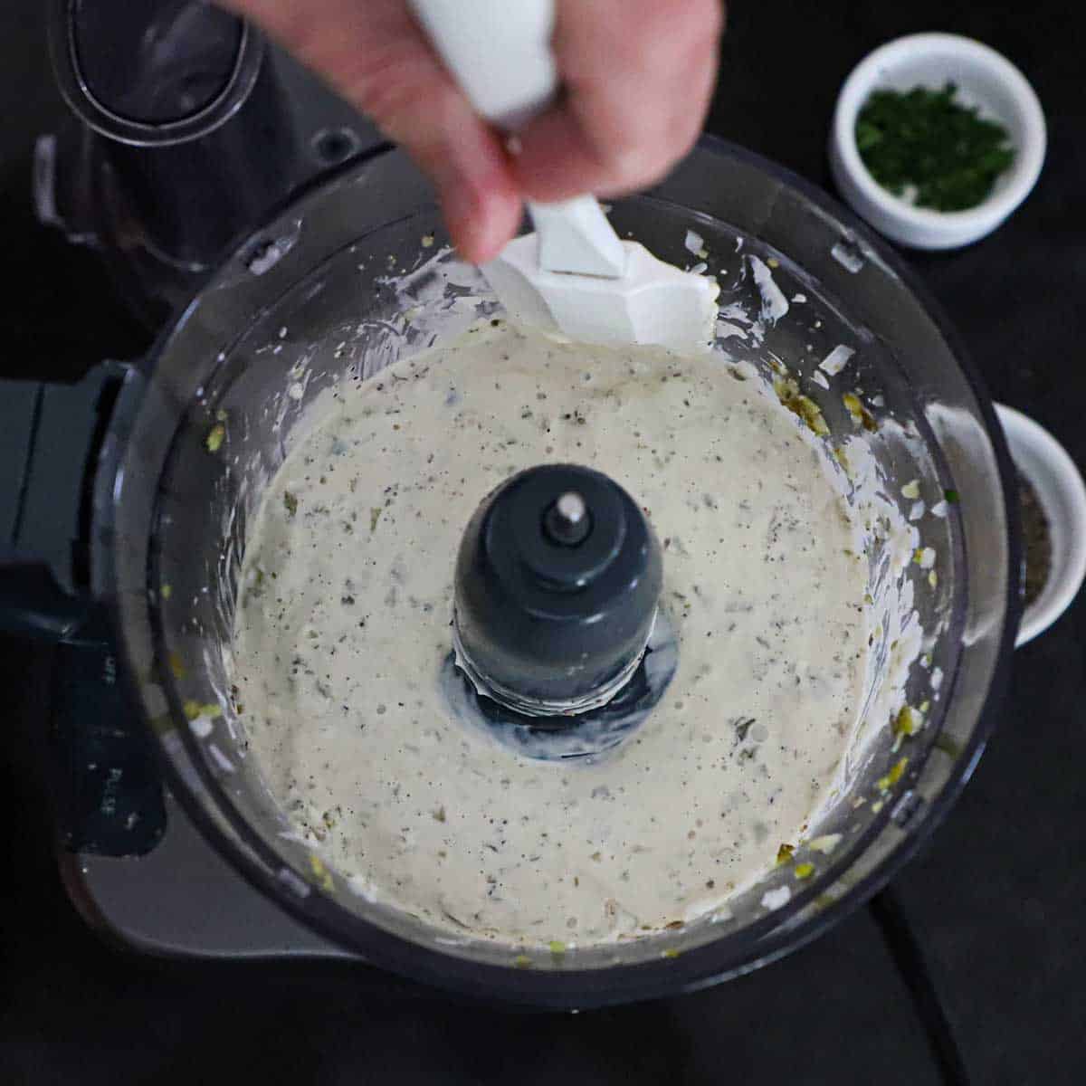 A person using a spatula to scrape down the sides of a food process bowl that has just blended the ingredients for homemade tartar sauce.