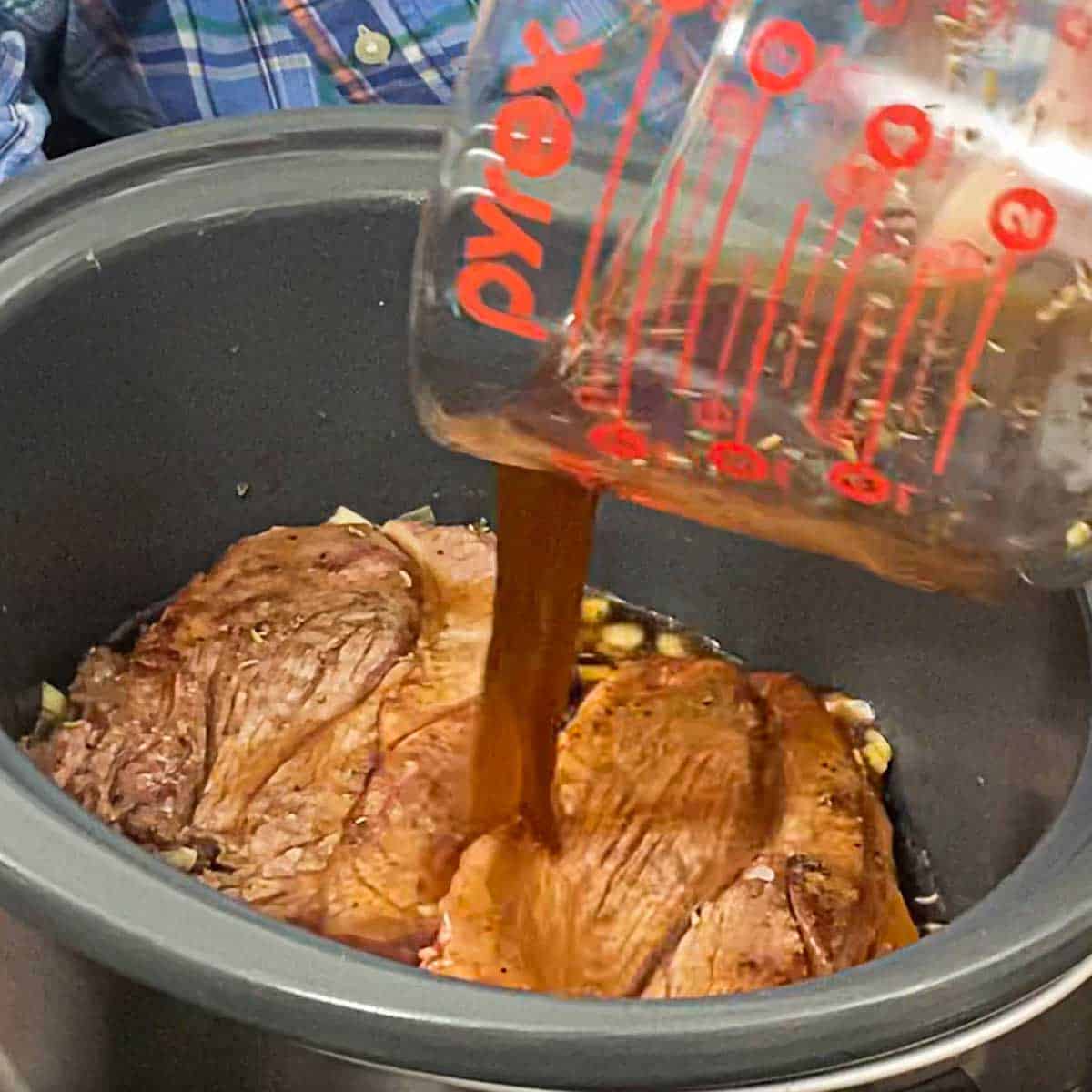 A person pouring beef broth from a large measuring cup over two seared and seasoned chuck roasts with chopped garlic in the bowl of a slow cooker.