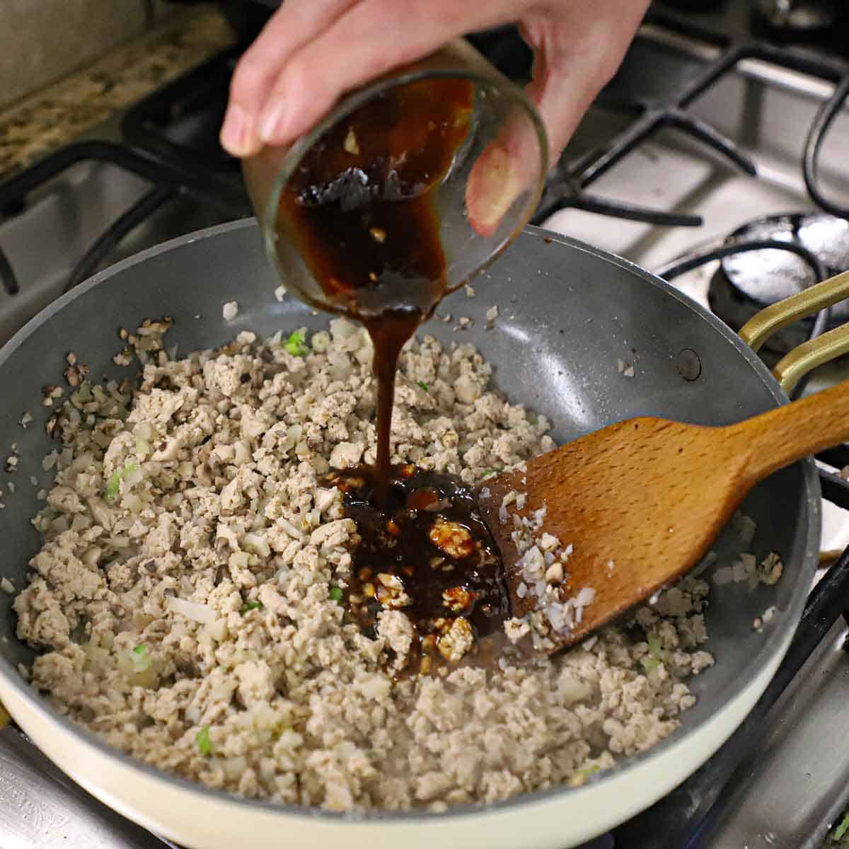 A person pouring a brown Chinese stir-fry sauce into a skillet that is filled with cooked ground chicken, chopped mushrooms, and chopped waterchestnuts.