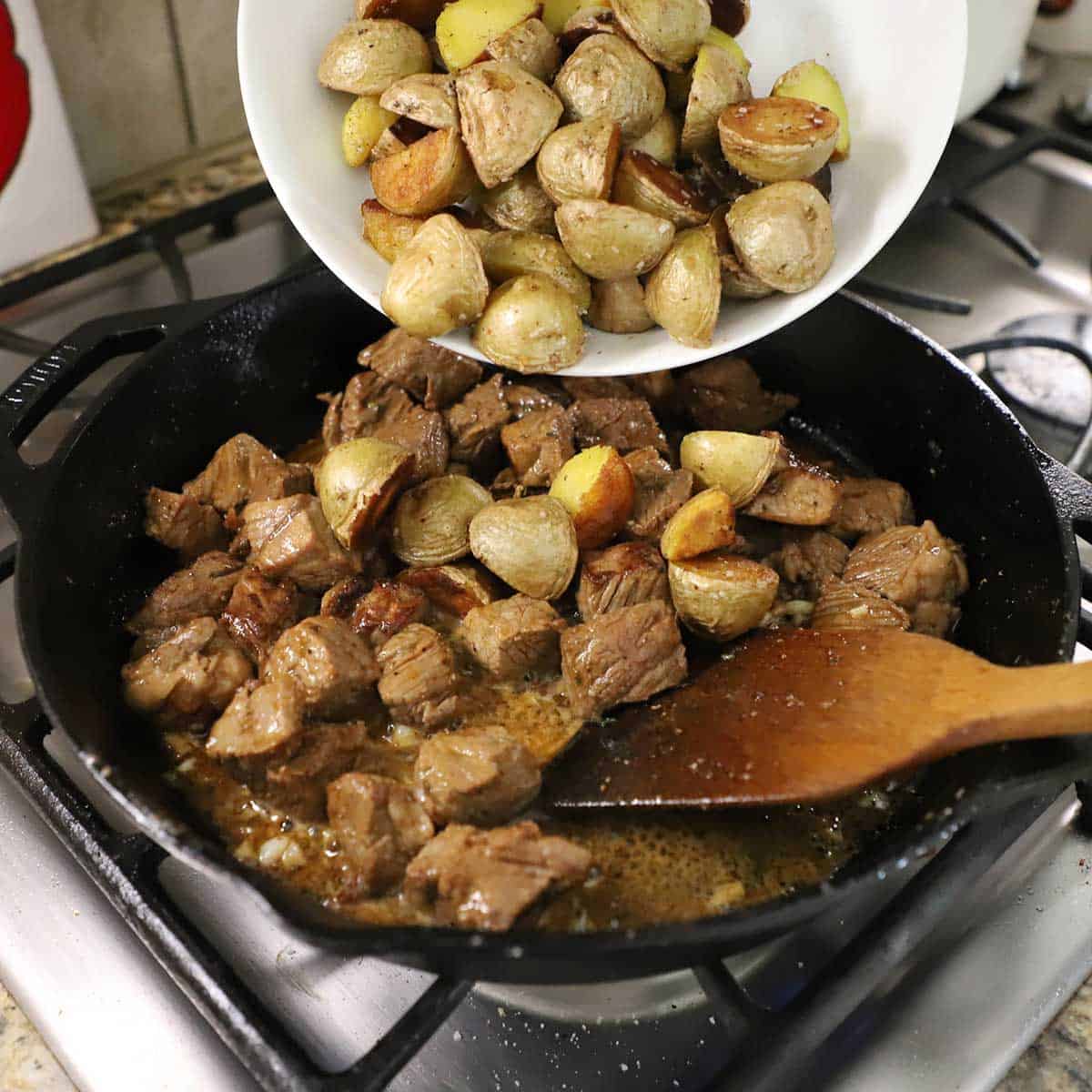 A person transferring seared halved baby gold potatoes from a white bowl into a skillet filled with garlic butter steak bites.