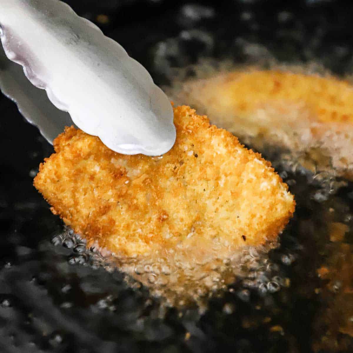 A person using a pair of metal tongs to turn a fried cod filet over in skillet filled with hot oil.