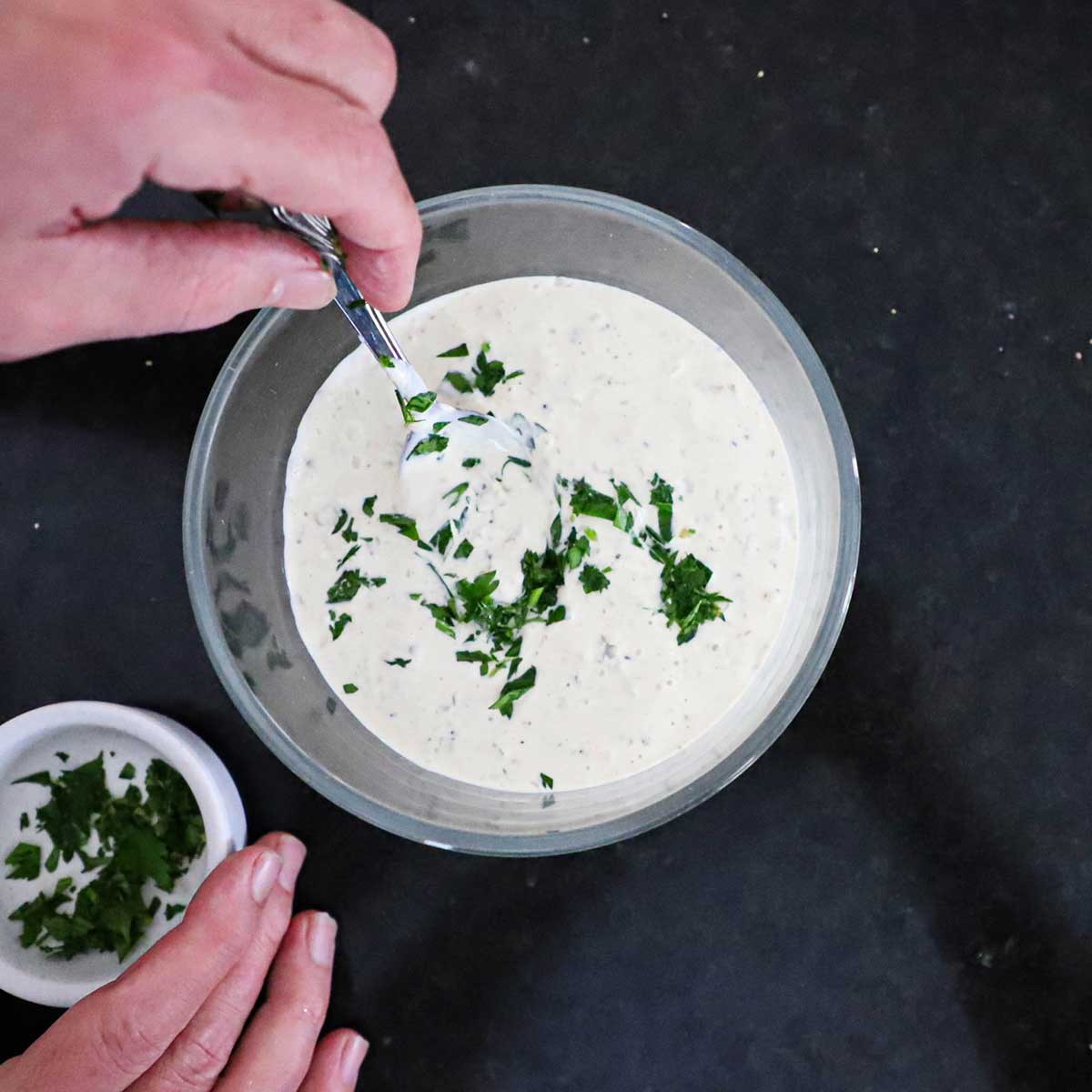 A person using a spoon to stir chopped Italian parsley into a small bowl filled with homemade tartar sauce.