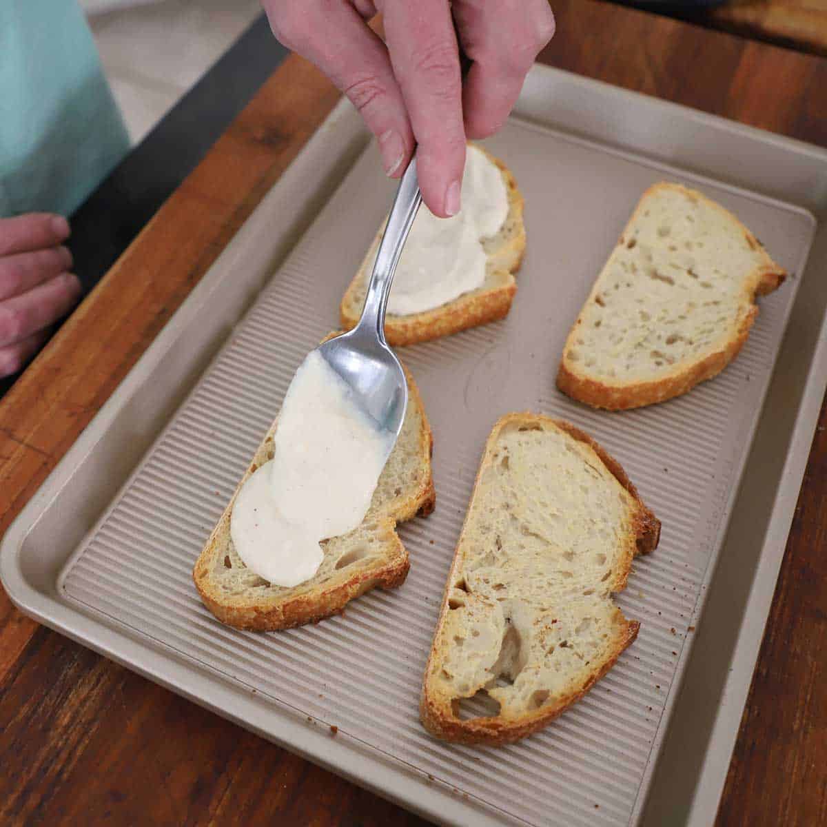 A person using a spoon to add a layer of bechamel sauce onto slices of toasted sourdough bread on a small baking pan on a wooden cutting board.