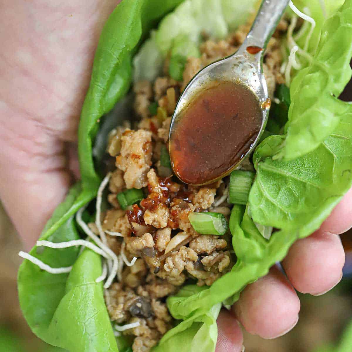 A person using a small spoon to add an Asian spicy sauce over the top of a copycat P.F. Change Chicken Lettuce Wrap that he is holding in his hand.