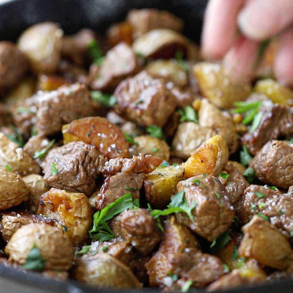 A person sprinkling chopped Italian parsley over the top of freshly prepared garlic butter steak bites and potatoes in a large cast-iron skillet.