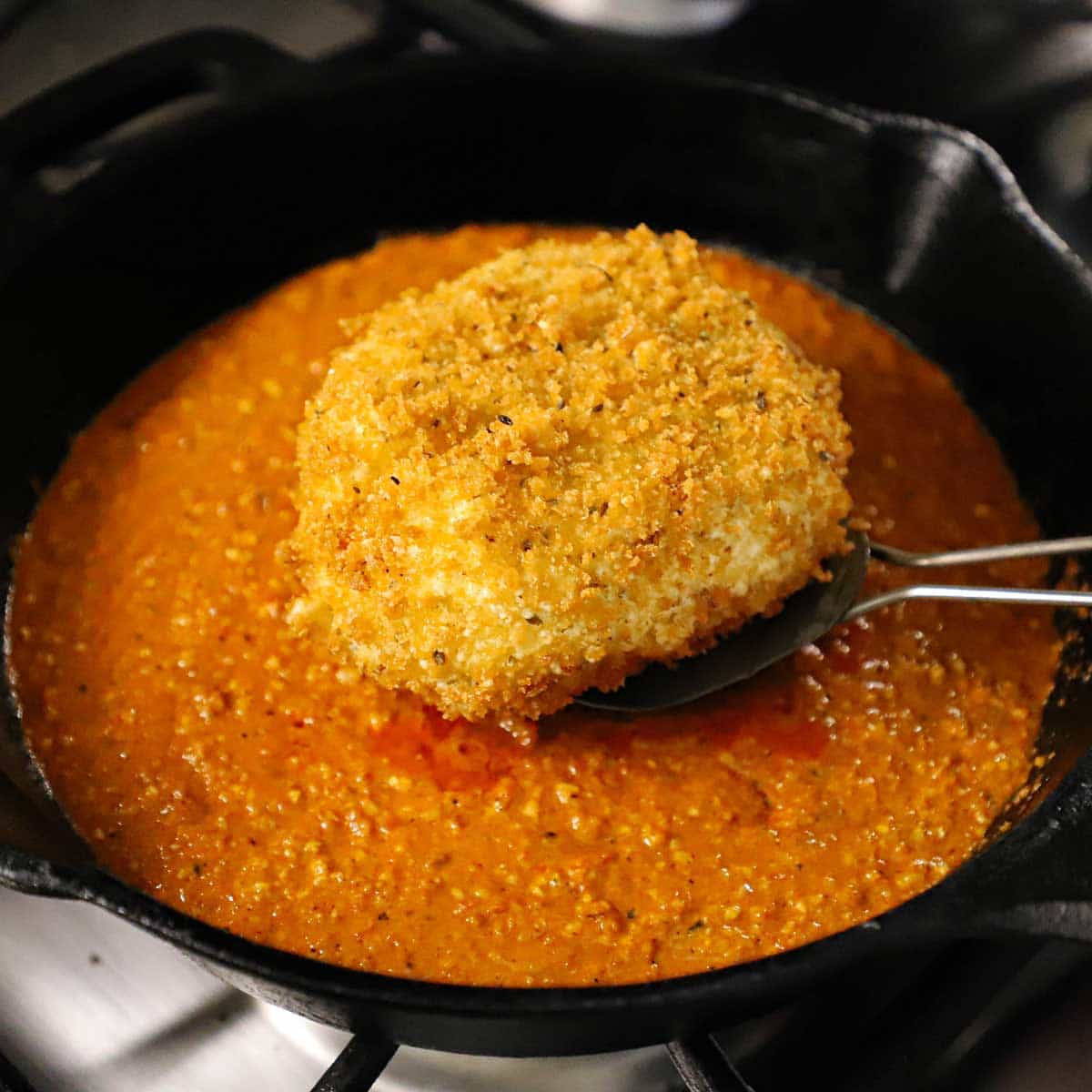 A person using a metal spatula to place a crispy fried burrata into a skillet that has a layer of smokey romesco sauce in it.