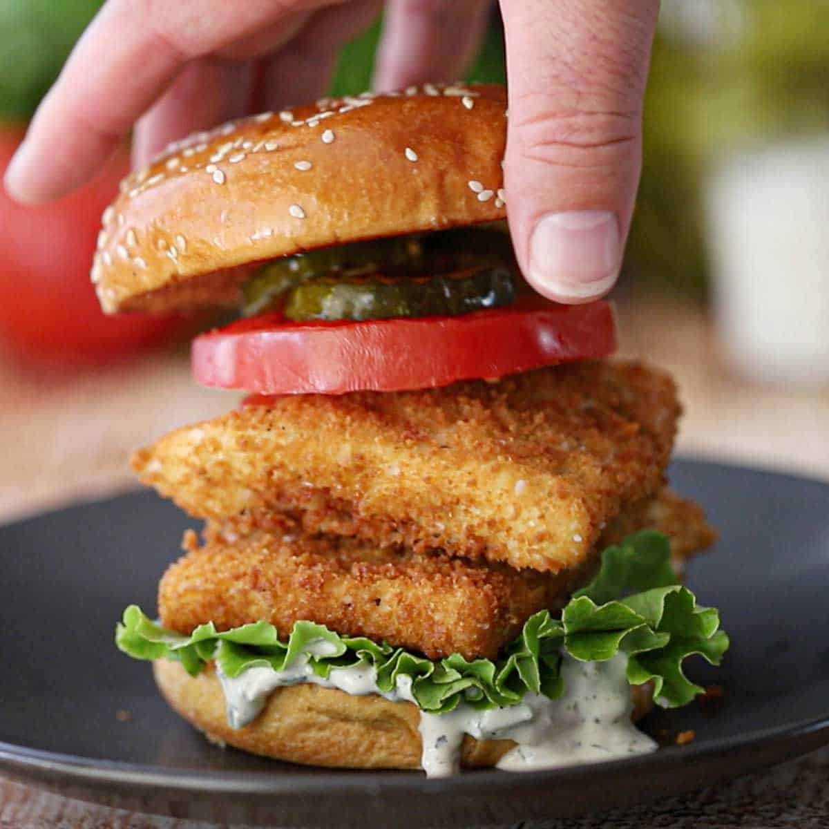 A person placing the top half of a toasted hamburger bun over the top of a fried fish sandwich that included to fried filets of cod, tartar sauce, lettuce, a tomato slice, and pickle chips.