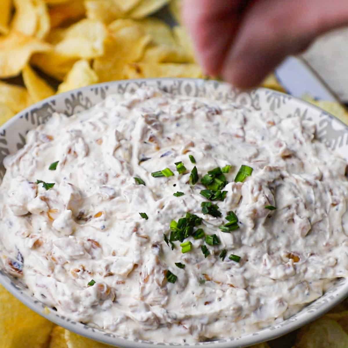 A person sprinkling snipped chives over the top of a small oval bowl that is filled with homemade French onion dip.
