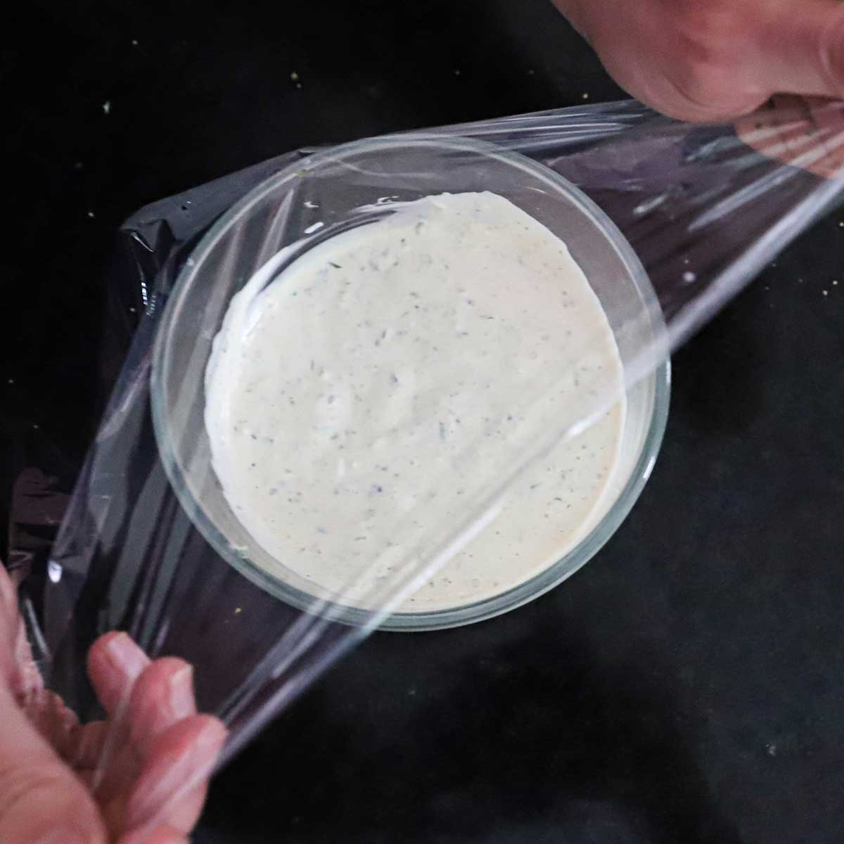 A person covering a small glass bowl filled with homemade tartar sauce with plastic wrap.