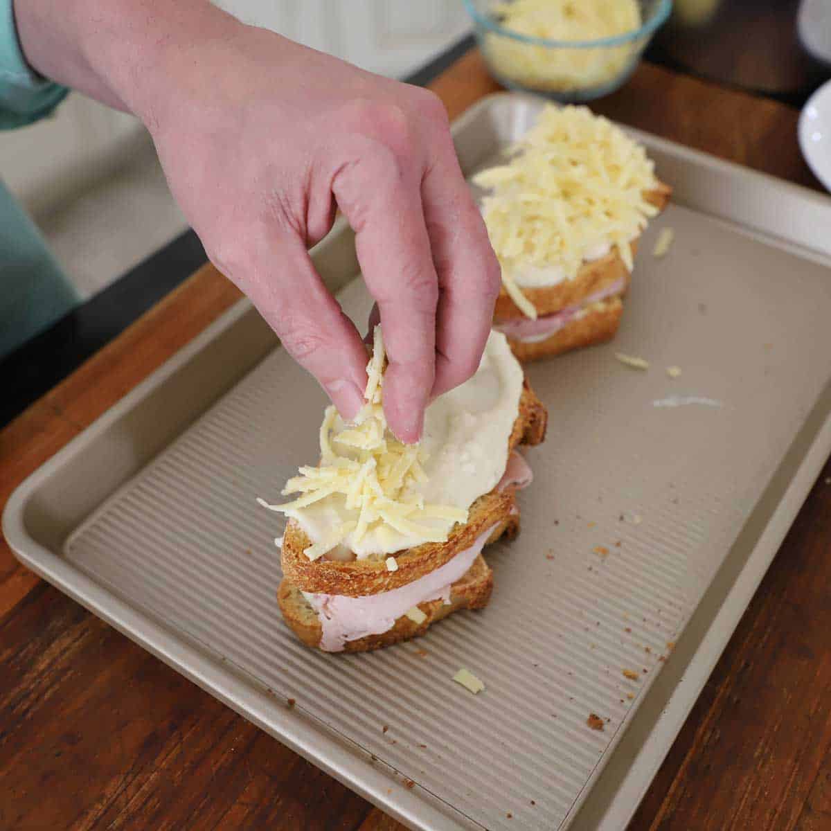 A person placing shredded Gruyere cheese over the top of assemble Essential Croque Monsieur sandwiches that are resting on a small baking pan.