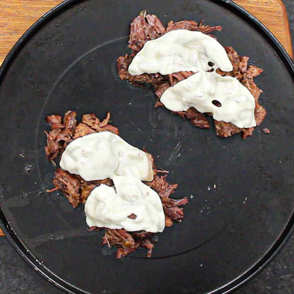 Two piles of shredded slow cooked beef on a round baking pan with melted provolone on top of the meat.