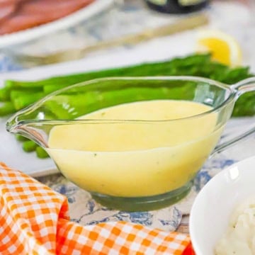 A close-up view of a glass gravy boat filled with Blender Béarnaise Sauce next to a platter of pan-seared asparagus.