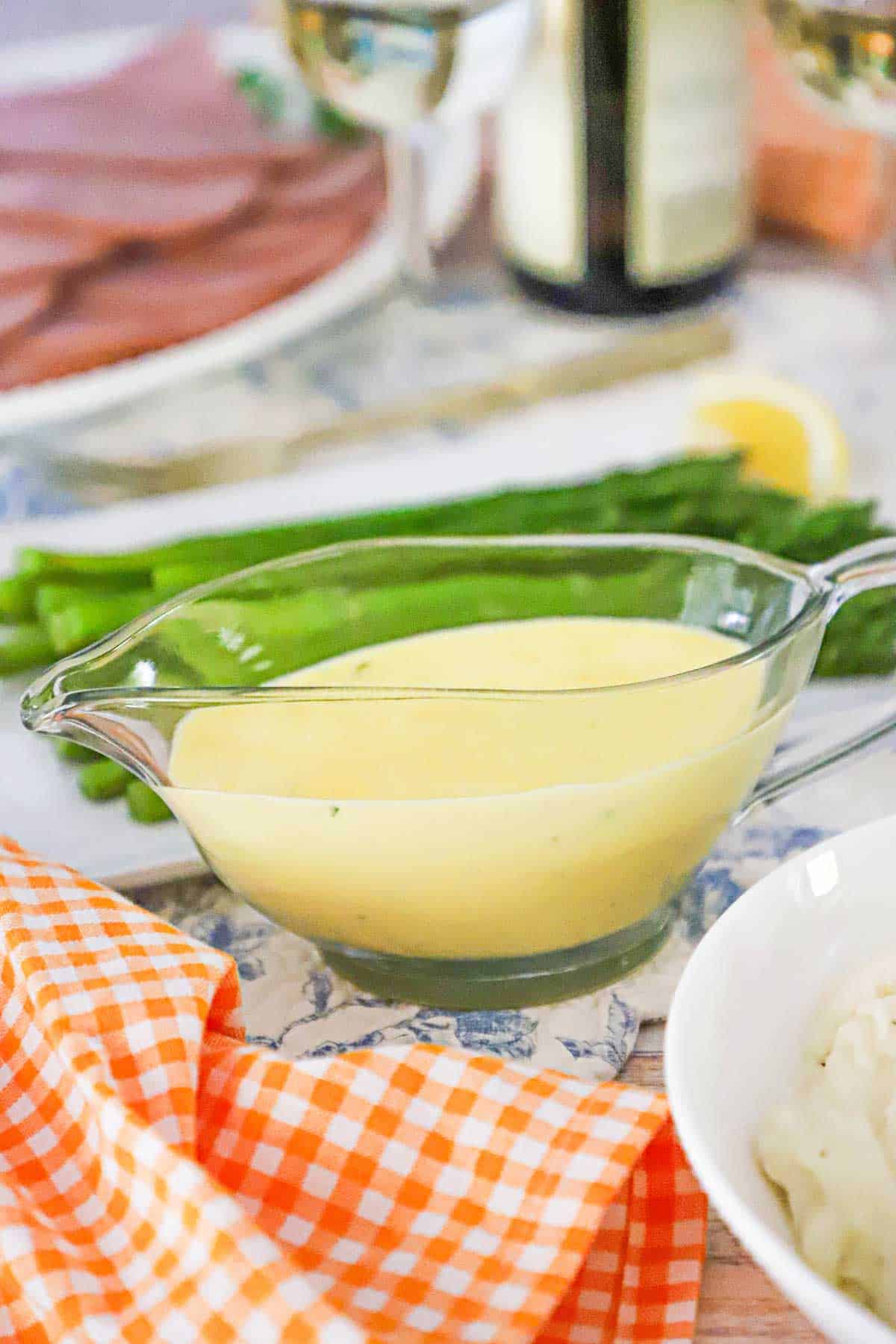 A glass gravy boat will with Blender B&eacute;arnaise Sauce next to an orange-checkered napkin and a platter filled with pan-seared asparagus.