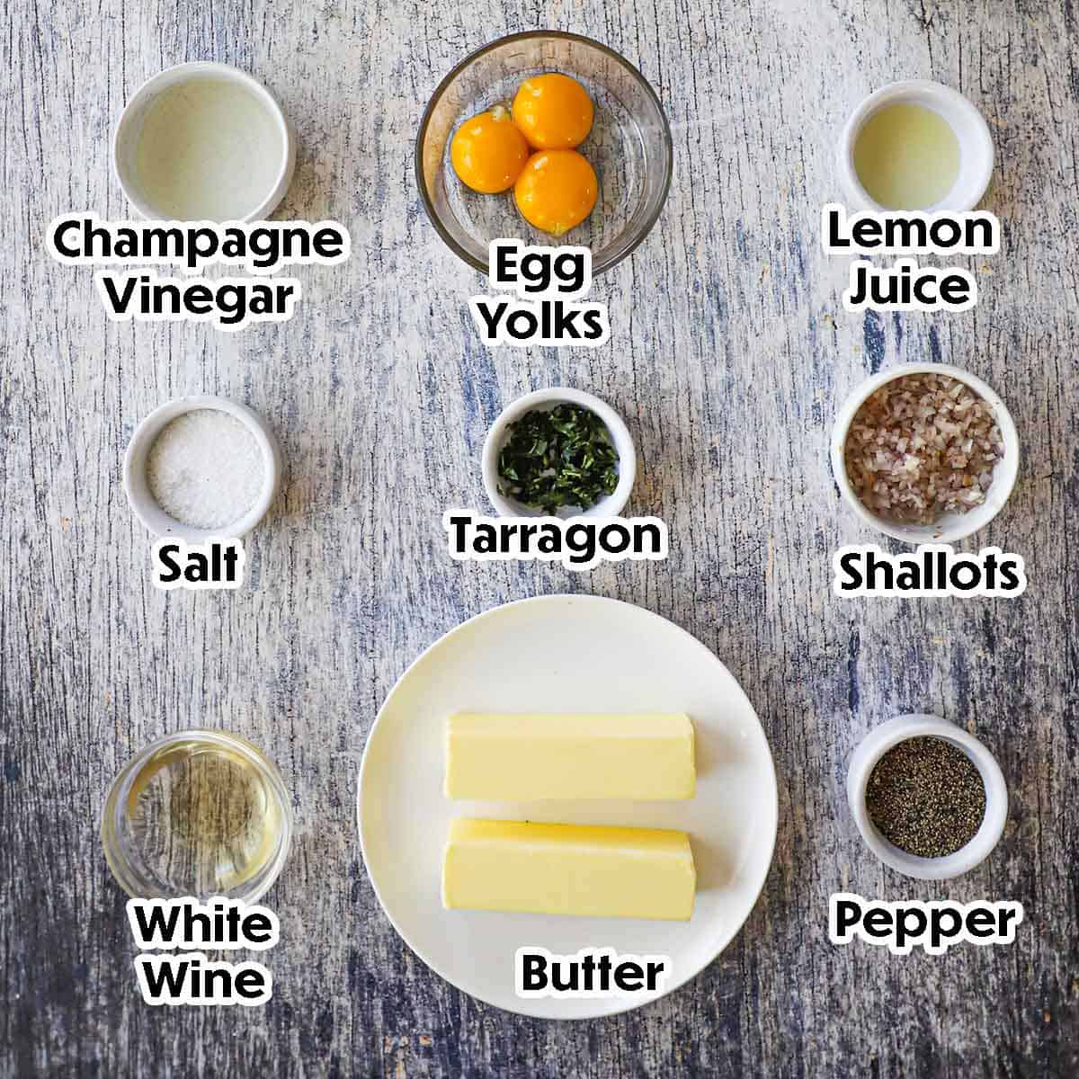 An arrangement of ingredients for Blender Béarnaise Sauce on a grey wooden background including two sticks of butter, egg yolks, vinegar, lemon juice, shallots, wine, tarragon, and seasonings.
