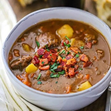 A bowl of classic Guinness beef stew in a white soup bowl and garnished with crispy bits of bacon and chopped Italian parsley.
