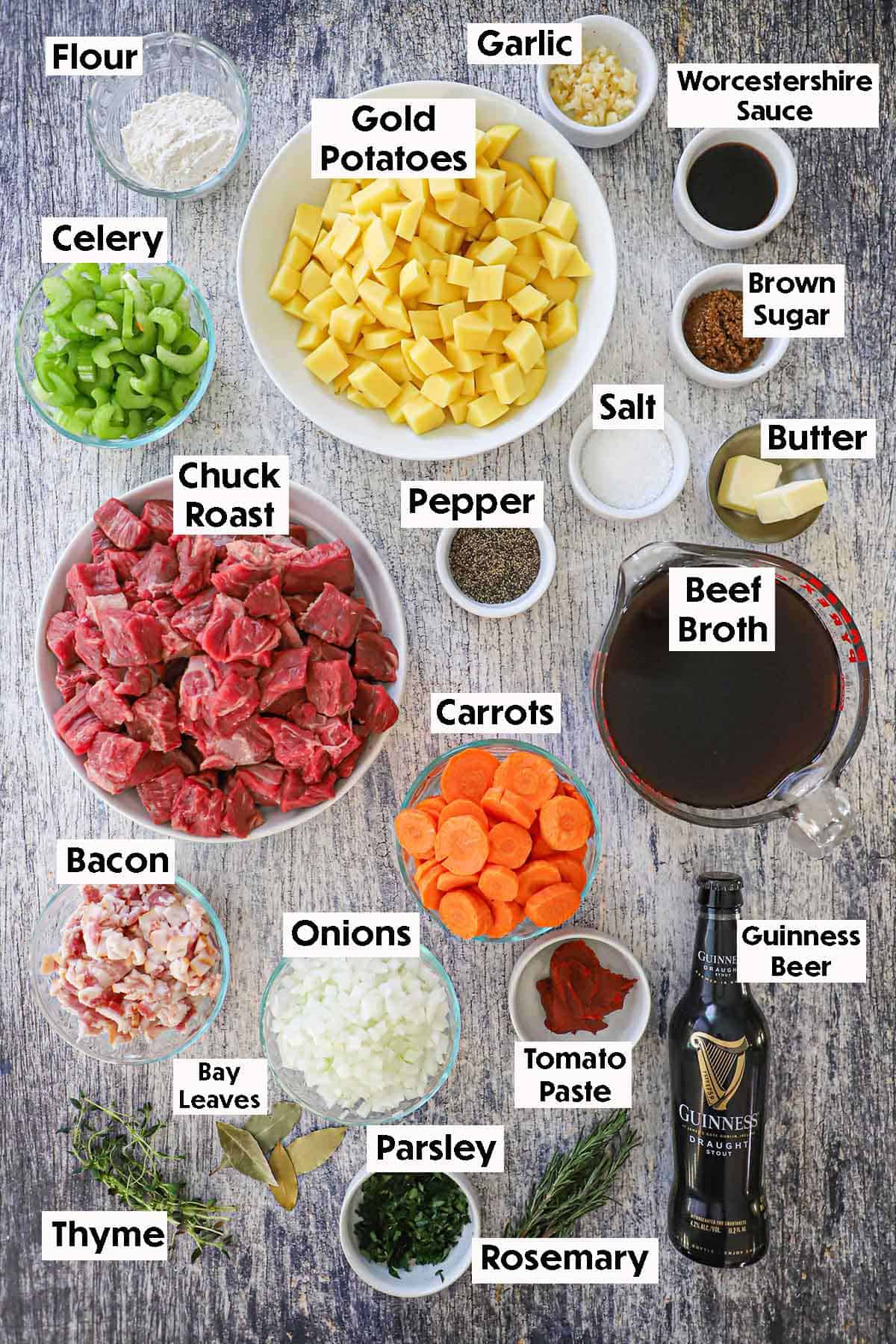 An arrangement of ingredients for Classic Guinness Beef Stew on a grey wooden background including chunks of chuck roast, potatoes, bacon, celery, carrots, garlic, beef broth, Guinness beer, brown sugar, and seasonings.