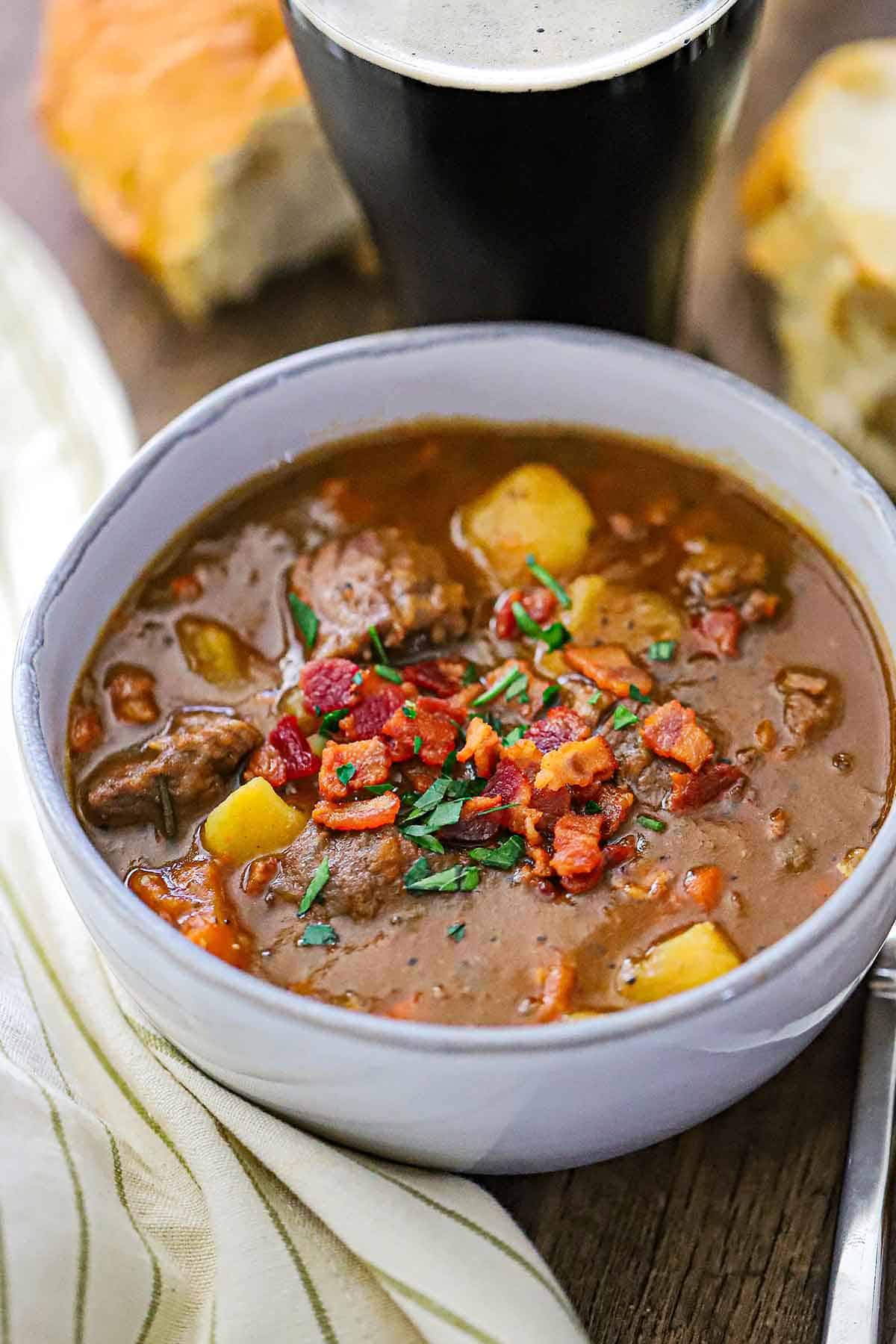 A bowl of classic Guinness beef stew in a white soup bowl and garnished with crispy bits of bacon and chopped Italian parsley.
