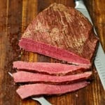 An overhead view of a homemade corned beef sitting on a wooden cutting board with several slices cut and showing the bright red hue of the meat.