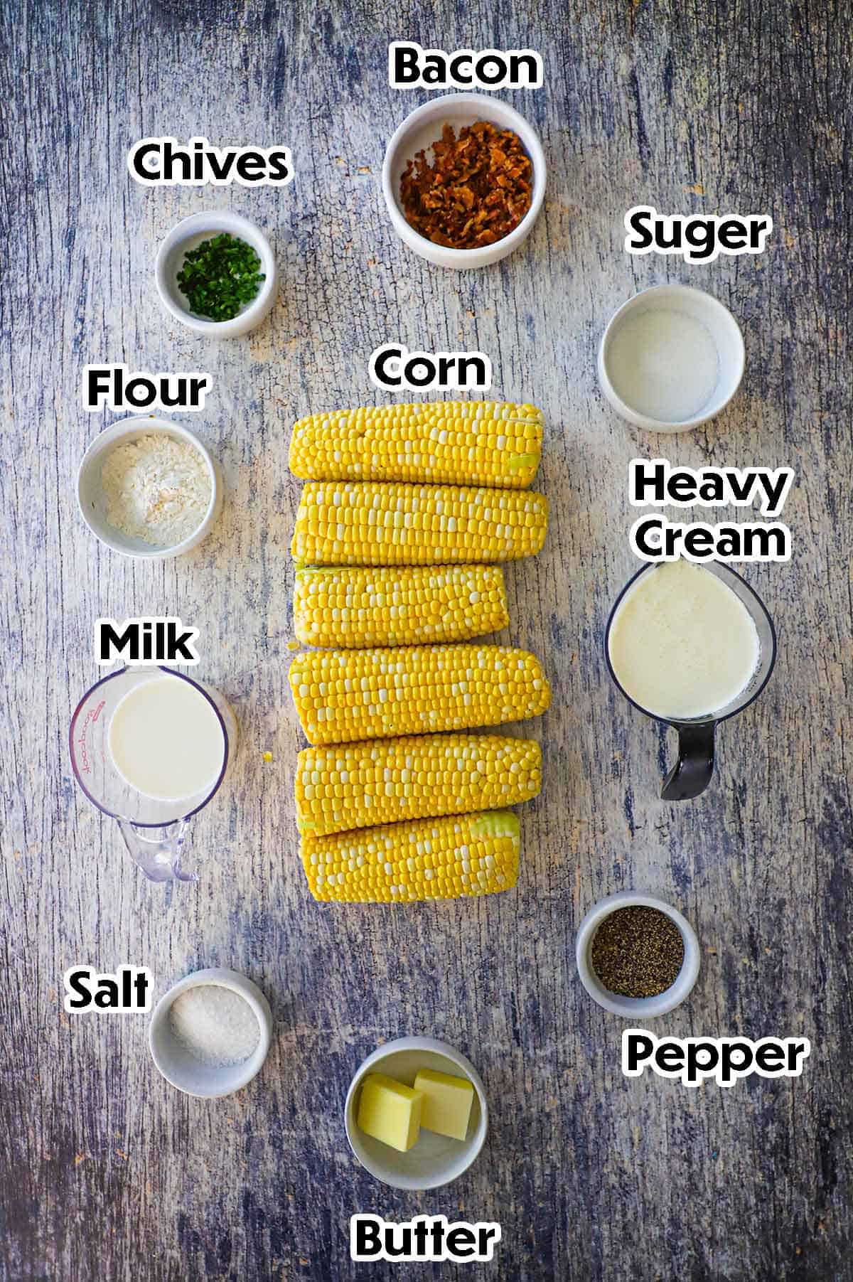 An arrangement of ingredients for homemade creamed corn on a grey wooden background including ears of corn, flour, sugar, cream, milk, bacon, chives, and seasonings.