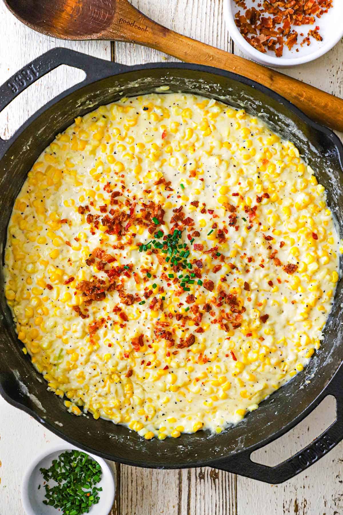 An overhead view a large cast-iron skillet with freshly prepared homemade creamed corn garnished with crispy bacon bits and snipped chives.