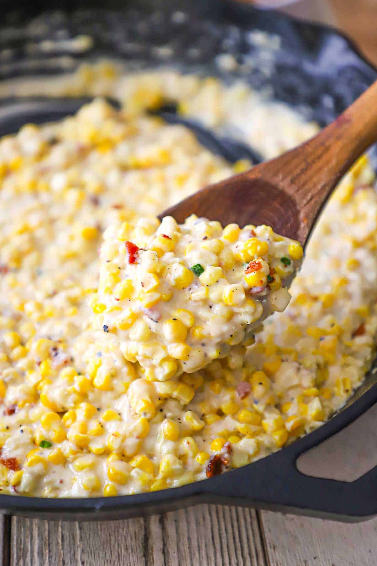 A close-up view of homemade creamed corn in a black cast-iron skillet with a wooden spoon being used to lift a serving of the corn from the pan.