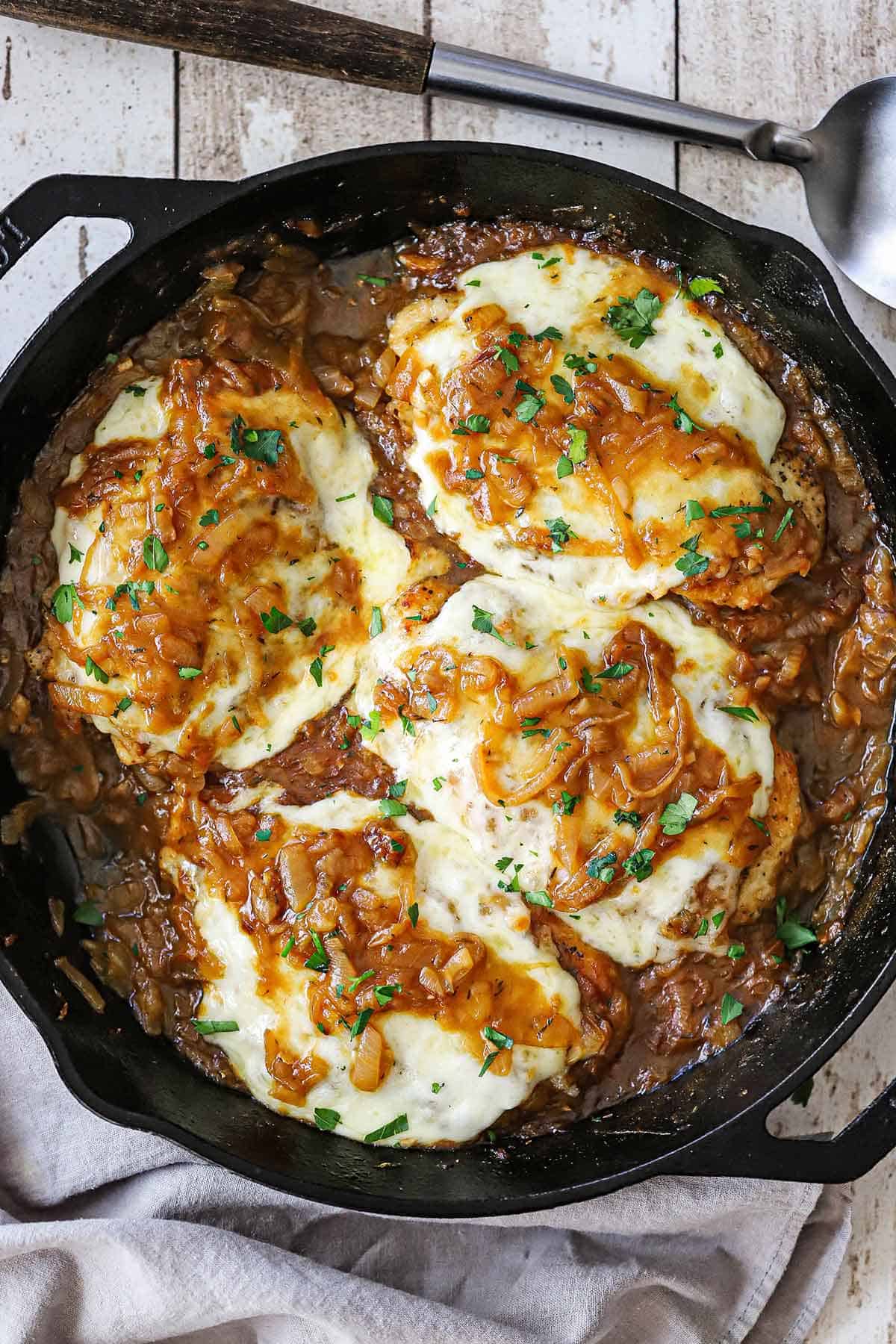 An overhead view of a large cast-iron skillet filled with four servings of French onion smothered chicken garnished with chopped Italian parsley.
