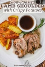An overhead view of a white dinner plate holding a serving of sliced slow-roasted lamb shoulder, crispy potatoes, and a bowl of red wine au jus.
