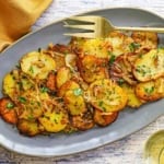 An overhead view of a grey, oval serving platter filled with a pile of freshly prepared Authentic Lyonnaise Potatoes with a large gold serving fork sitting on the edge of the platter.