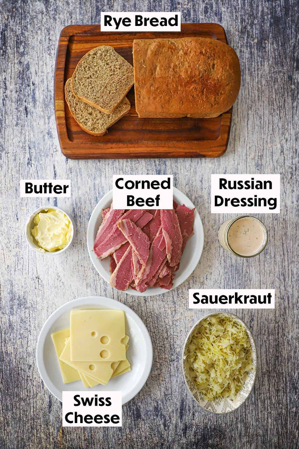 An overhead view of the ingredients for "From Scratch" Reuben Sandwich including a pile of corned beef, rye bread, Swiss cheese, sauerkraut, Russian dressing, and softened butter.
