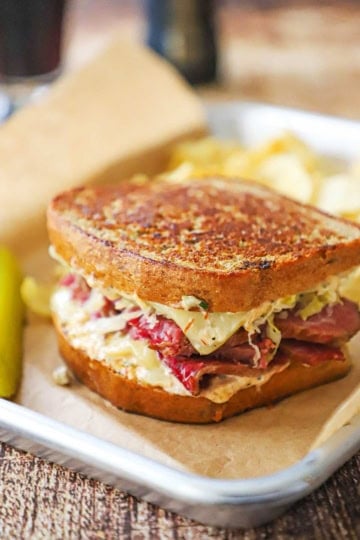 A close-up view of a "From Scratch" Reuben Sandwich resting on a brown paper in a small metal sheet pan with a pickle spear and kettle chips nearby.