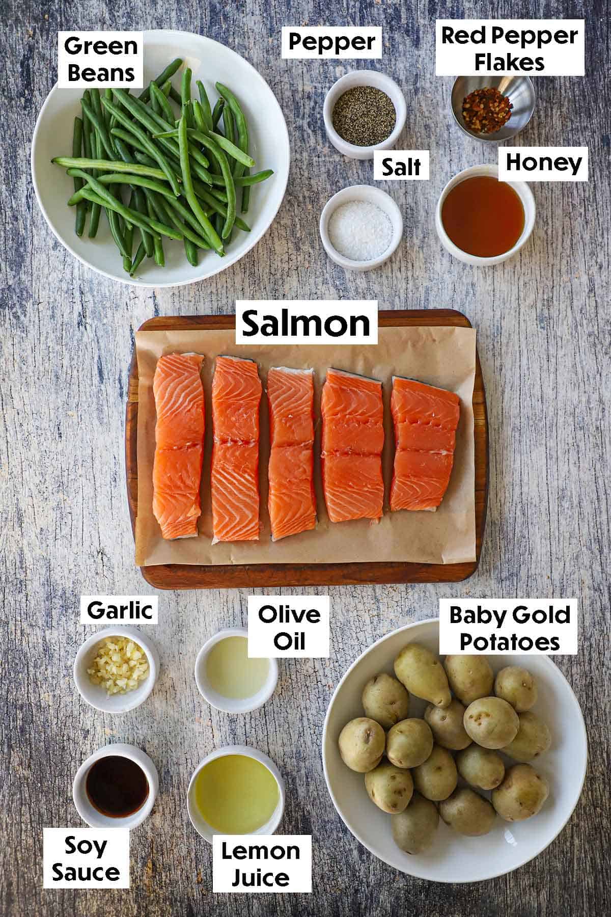 An arrangement of ingredients for sheet pan honey garlic salmon and roasted vegetables on a wooden grey background including salmon filets, green beans, baby gold potatoes, garlic, honey, lemon juice, and seasonings.