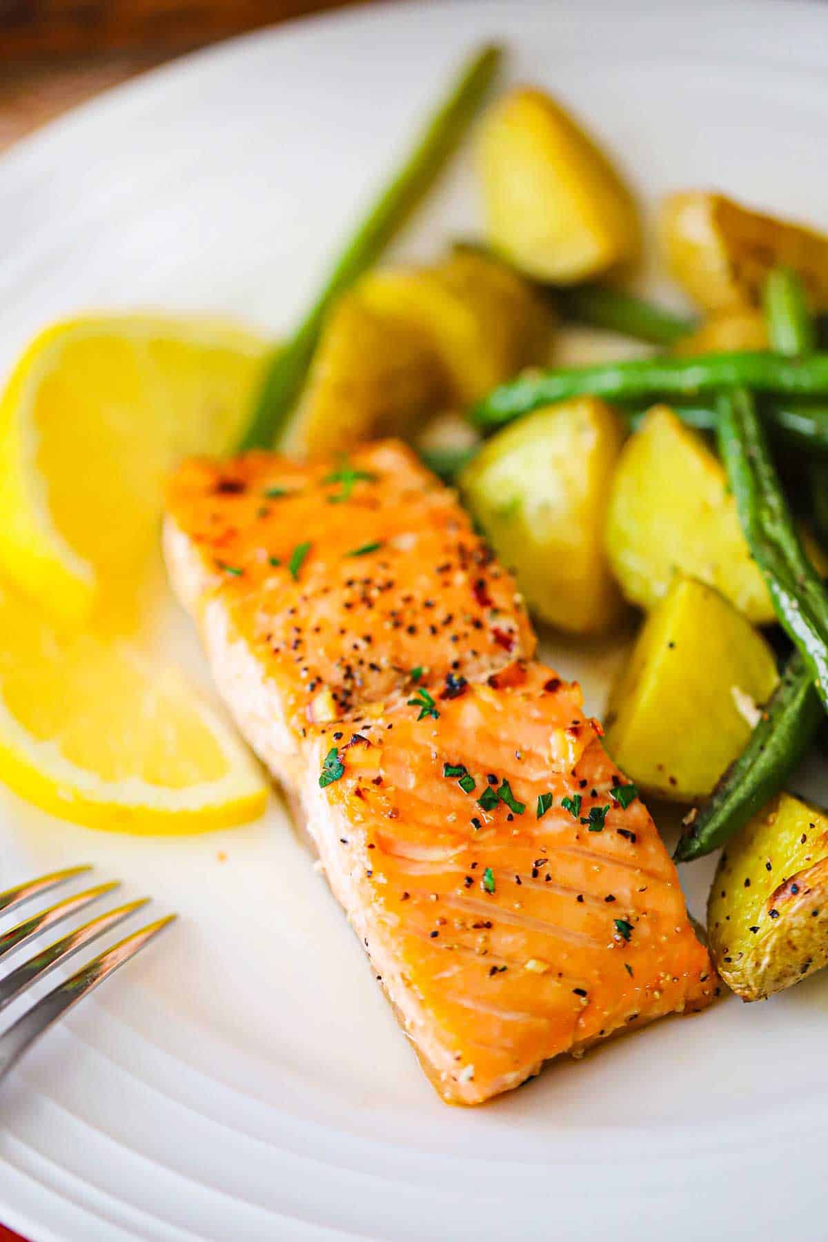 A close-up view of a fillet of roasted honey garlic salmon next to oven-roasted potatoes and green beans and a couple slices of lemon.