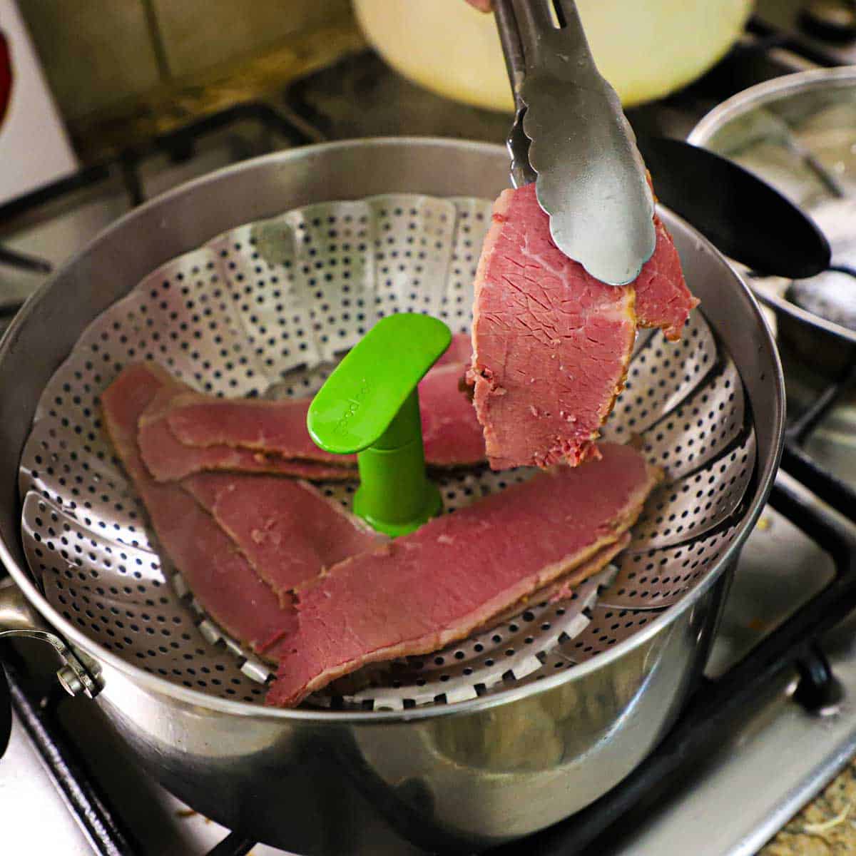 A person placing thin slices of cooked corned beef into the basket of a steamer that is nestled into a pot with simmering water.