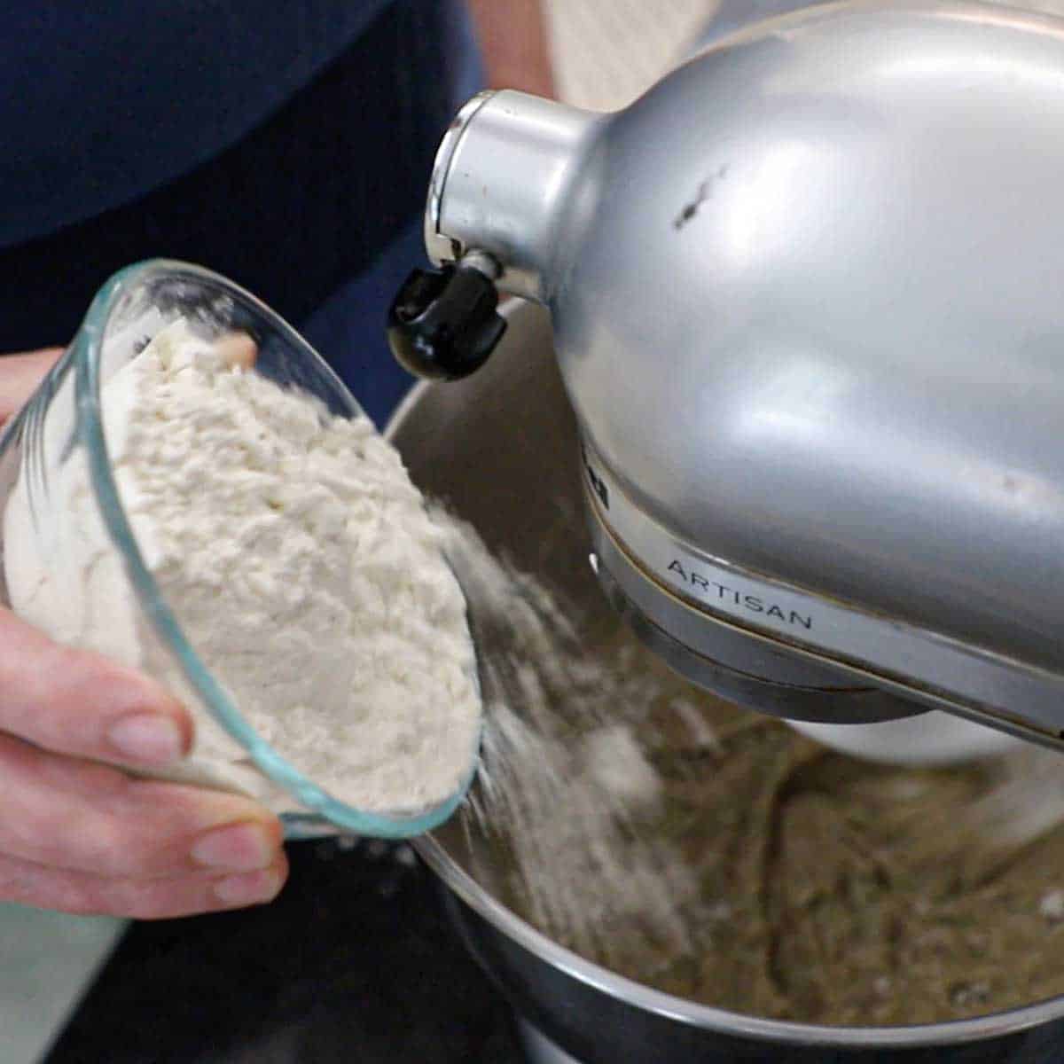 A person adding rye floured into a stand-mixer that contains yeast that has been activated in a warm water and honey.