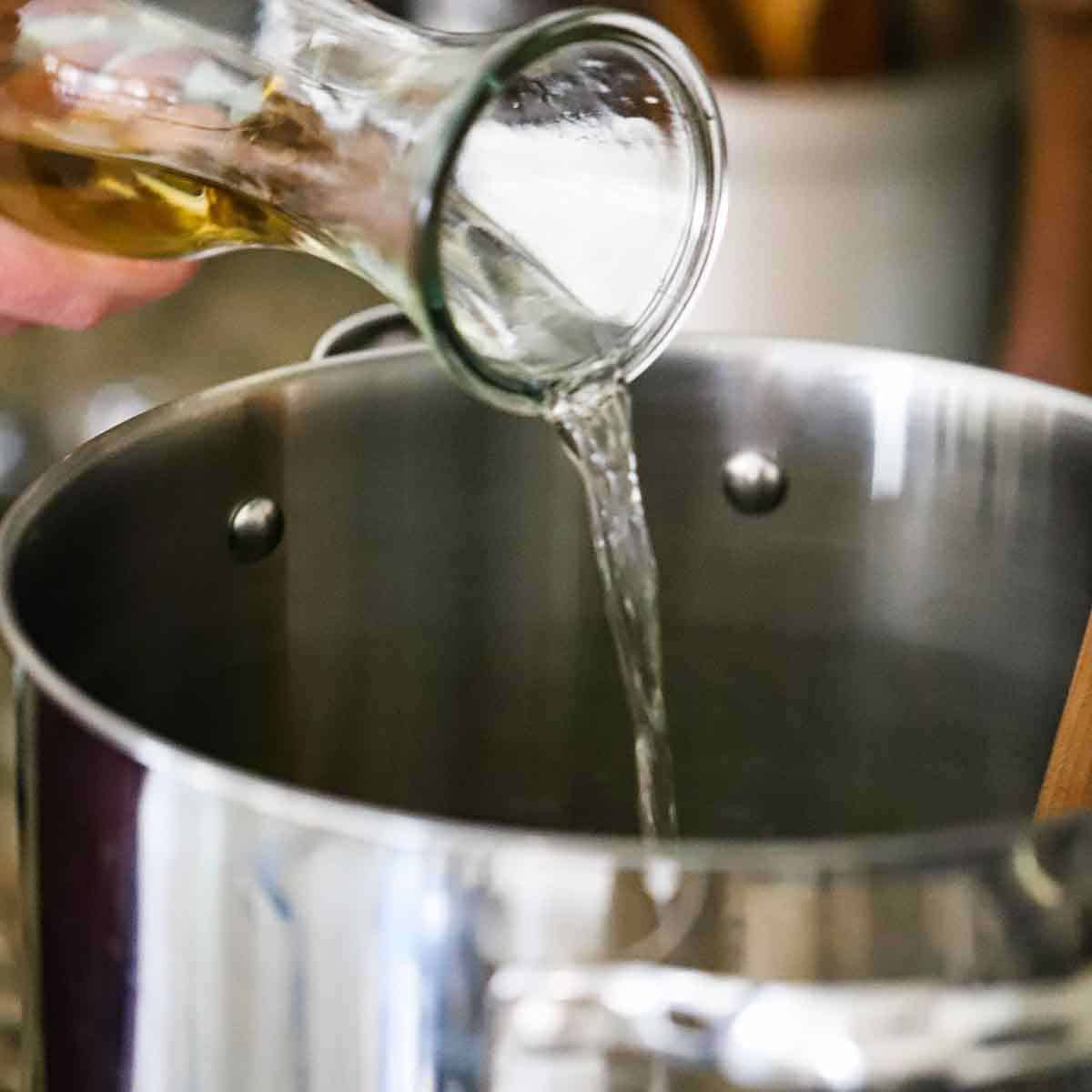 A person pouring white wine from a carafe into a large stock pot with a wooden spoon resting in it.