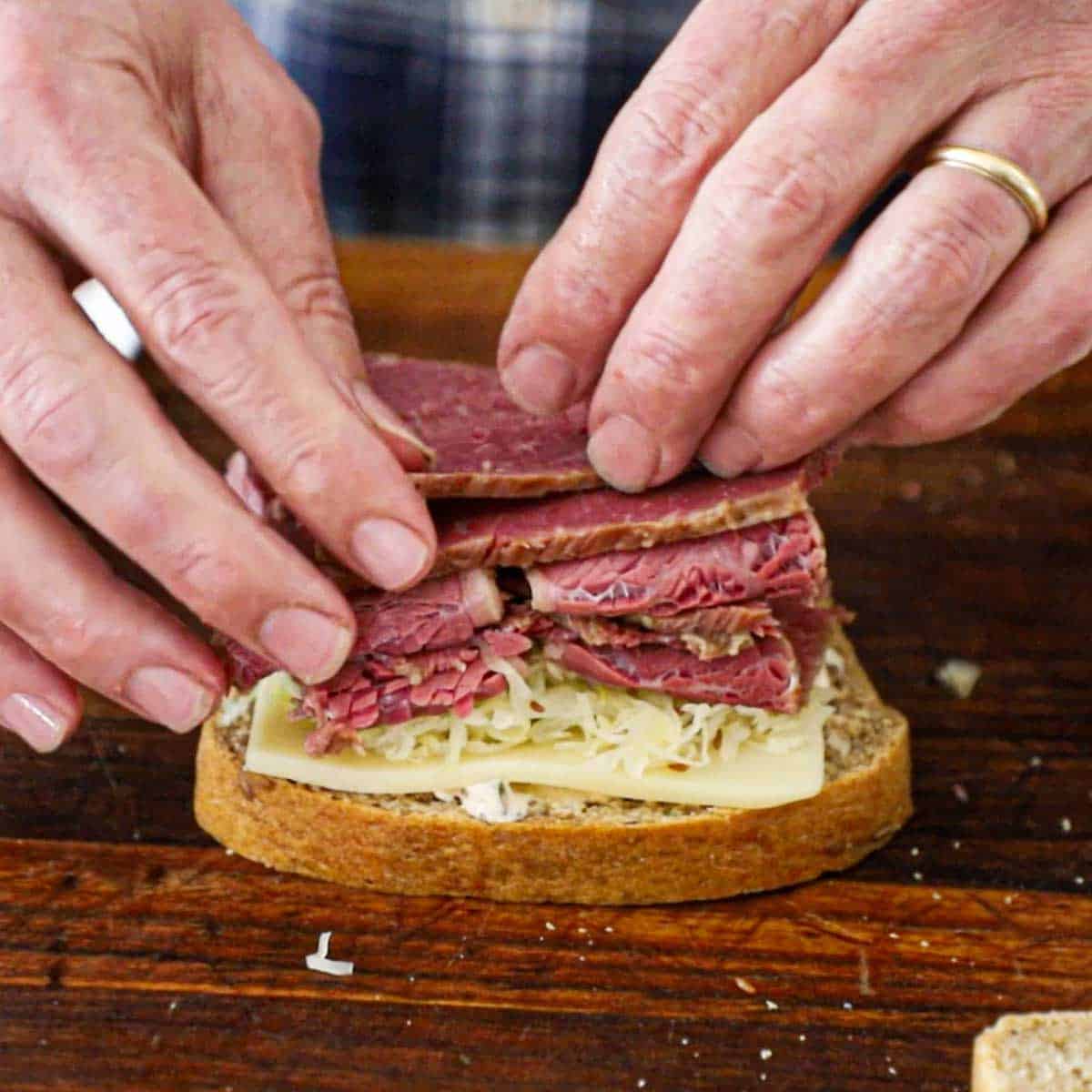 A person pile warm, thin slices of corned beef over the top of sauerkraut resting on top of Swiss cheese over a smear of Russian dressing on a slice of homemade rye bread.