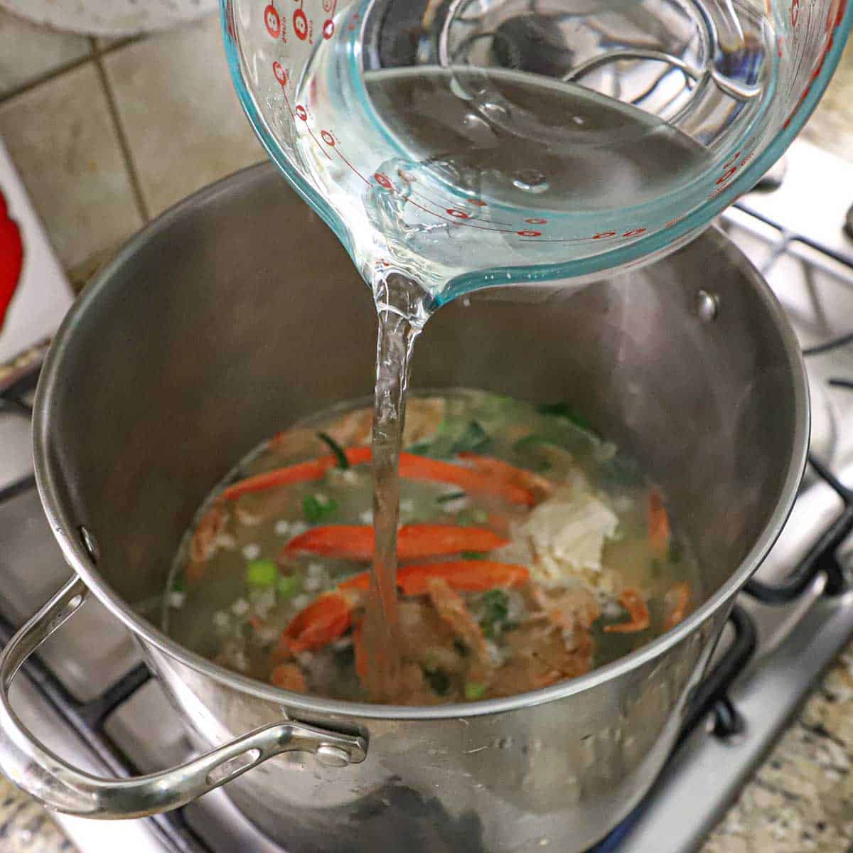 A person pouring water from a large 10-cup glass measuring cup into a large stock pot containing the ingredients for homemade shellfish stock.