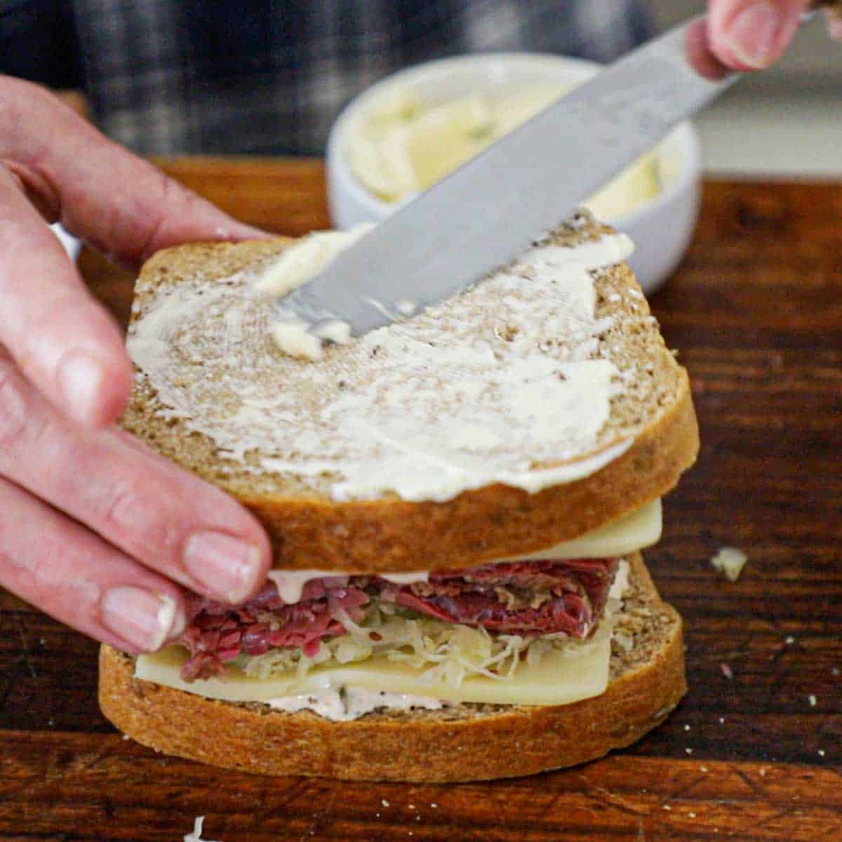 A person smearing softened butter over the top slice of a "from scratch" reuben sandwich that is about to be griddled.